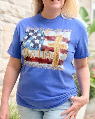 Person wearing a blue t-shirt with a graphic design featuring an American flag and cross.