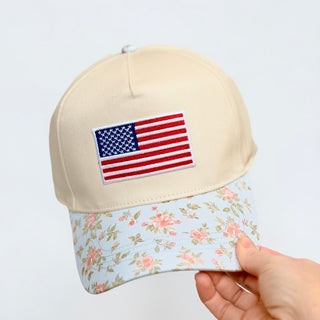 Beige cap with American flag patch and floral brim held by a hand on a light gray background