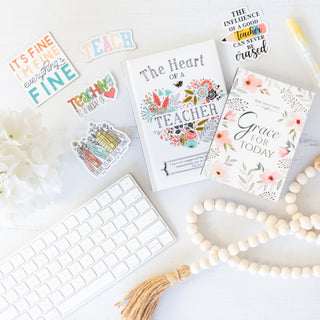 Desk flat lay with teacher appreciation stickers, devotional books, keyboard, beaded garland, and pen on white surface