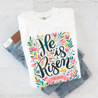 White graphic tee with colorful floral design and "He is Risen" text, paired with light blue denim jeans.