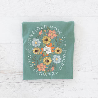 Consider How the Wild Flowers Grow Luke 12:27-28 graphic on a folded sage green t-shirt