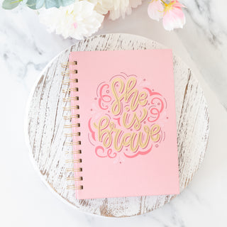 She is Brave spiral wire bound pink journal on a white wooden board with flowers