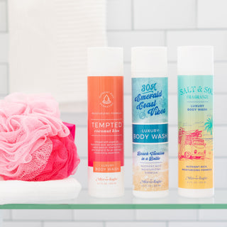 Three Mix-o-logie luxury body wash bottles and a pink shower pouf on a glass shelf