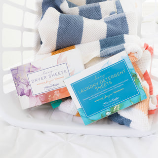 Mixologie dryer sheets and laundry detergent sheets in a laundry basket with striped towels