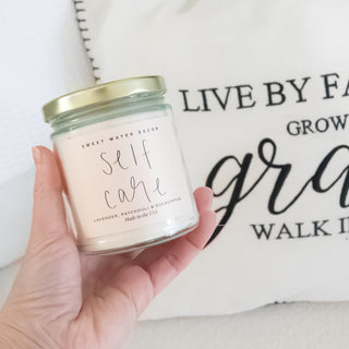 Hand holding Self Care Scented Soy Candle by Sweet Water Decor with Christian faith-based decorative pillow background