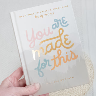 Hand holding You Are Made for This Devotions to Uplift & Encourage Busy Moms book with soft pastel cover and gold lettering.