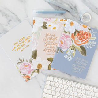 Three floral notebooks with scripture verses on a marble desk with pens and a keyboard