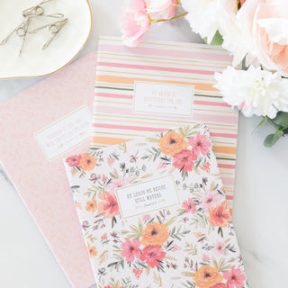 Three floral and striped notebooks with scripture verses on a desk with flowers