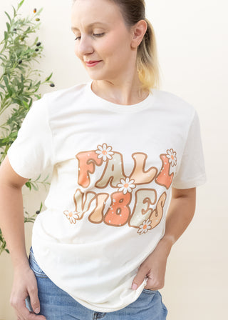 Person wearing a cream color graphic tee with Fall Vibes text and retro daisy illustrations