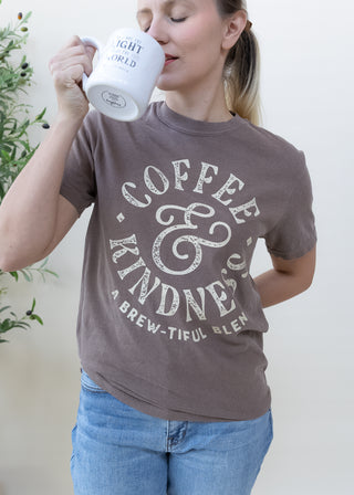 Woman wearing brown Coffee and Kindness A Brew-tiful Blend graphic t-shirt while drinking from a white mug