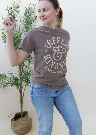 Woman wearing a brown Coffee and Kindness A Brew-tiful Blend graphic t-shirt with blue jeans