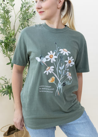Woman wearing dark green Rejoice in Hope Be Patient in Trials Keep on Praying graphic tee with daisy illustration