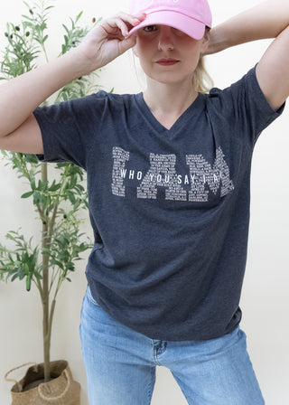 Person wearing I Am Who You Say I Am graphic tee in heather black with pink cap and blue jeans