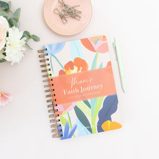 Moms Faith Journey A Year of Reflections and Testimony spiral notebook with floral cover and pen on white surface