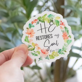 Hand holding He Restores My Soul Psalm 23:3 Wreath Sticker with floral wreath and green leaves background
