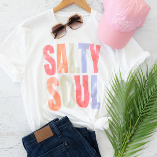 White t-shirt with 'Salty Soul' text, pink cap, sunglasses, and jeans on a light wooden surface.