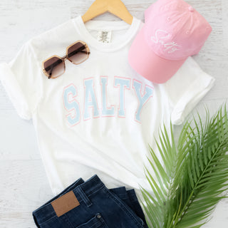 White t-shirt with 'Salty' text, pink cap, sunglasses, and jeans on a light gray background.