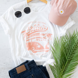 White t-shirt with beach-themed design, pink cap, sunglasses, and jeans on a light wooden surface.