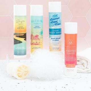Four bottles of Mixologie Luxury Body Wash with a loofah and towel on a white surface