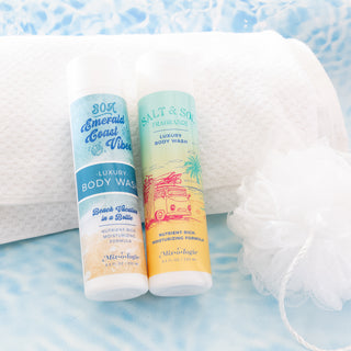 Two Mixologie luxury body wash bottles with a white towel and bath pouf on a blue background