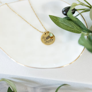 Gold necklace with Sunday Olive pendant and green gemstone on a white plate with olive branches