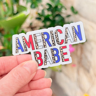 Sticker with 'AMERICAN BABE' text held by a hand against a blurred natural background