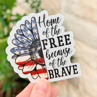 Sticker with American flag design and text 'Home of the Free because of the Brave' held by a hand.