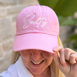 Woman wearing Salty in Script Writing Bright Pink Baseball Cap smiling and adjusting it outdoors with greenery background