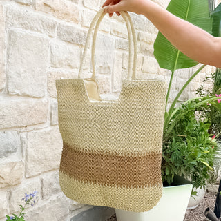 Woven straw bag held by a hand against a stone wall and plant background