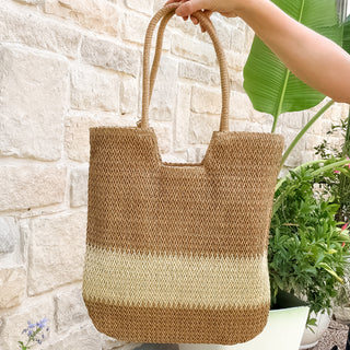 Woven tote bag held by a hand against a stone wall with plants.