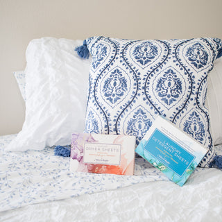 Mixologie luxury dryer sheets and laundry detergent sheets boxes on a bed with patterned pillows